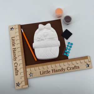 May include: A white ceramic Easter basket craft kit with a bow, a paintbrush, paint pots, and blue gems. The kit includes a brown felt square and wooden rulers with the text "Little Handy Crafts".