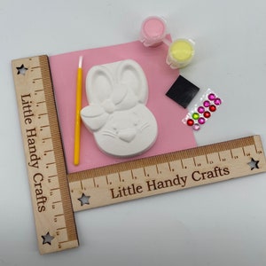 May include: A craft kit with a white bunny-shaped plaster mold, paint pots, a paintbrush, and decorative gems. The kit includes a pink mat and wooden rulers with the text "Little Handy Crafts".