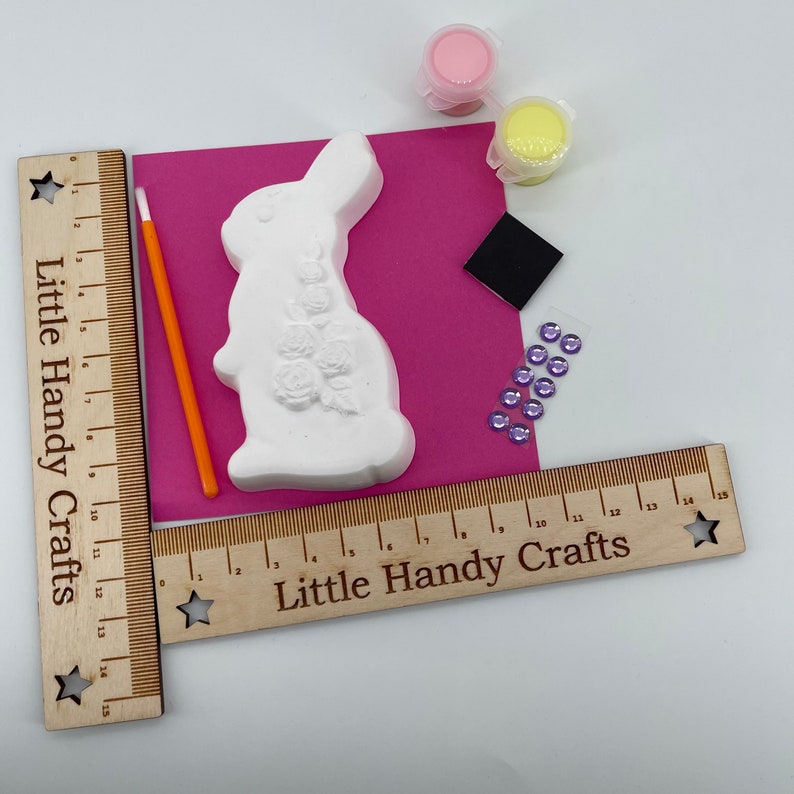 May include: A white plaster bunny figurine with rose detailing, paint pots, a paintbrush, and embellishments. The kit includes a pink background and wooden rulers with the text "Little Handy Crafts".