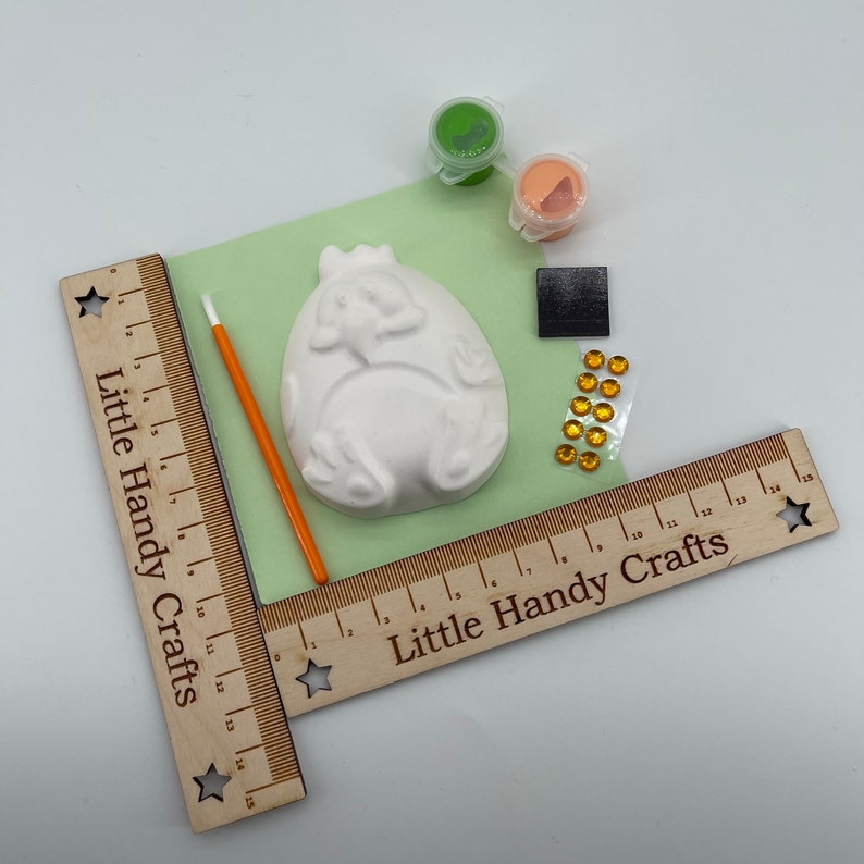 May include: A craft kit with a white ceramic egg-shaped figure with an embossed design, paint pots, a paintbrush, and decorative gems. The kit includes a wooden ruler with the text "Little Handy Crafts".
