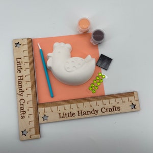 May include: A craft kit featuring a white ceramic chicken, paint pots, a paintbrush, and decorative gems. The kit includes a wooden ruler with the text "Little Handy Crafts". The chicken is on a peach-colored paper.