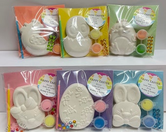 May include: Easter-themed craft kits featuring ceramic figurines of a chick, bunny, and egg, each with paint pots, a brush, and decorative gems. The kits are packaged in clear plastic bags with colorful backing cards that read "Happy Easter".