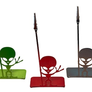 May include: Three metal alien-shaped memo holders in green, red, and gray. Each has a rectangular base and a tall, thin stem with a clip at the top. The alien figures have large heads and outstretched arms.