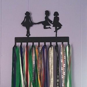 May include: Black metal medal hanger with three silhouette figures of dancers holding hands. The hanger has hooks for hanging medals and ribbons. The ribbons have text that says "2015" and "THIRD".