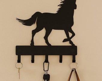 May include: Black metal key holder with a horse silhouette design. The key holder has five hooks and is mounted on a beige wall. A brown leather tote bag hangs from one of the hooks. A wooden table and a woven basket are also visible.