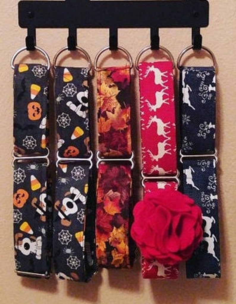 K&ouml;nnte beinhalten: F&uuml;nf verschiedene gemusterte Hundehalsb&auml;nder h&auml;ngen an einem schwarzen Metallgestell. Die Halsb&auml;nder sind aus Stoff und haben verschiedene Designs, darunter Mais, Spinnen, Bl&auml;tter, Rentiere und ein Schneeflockenmuster.