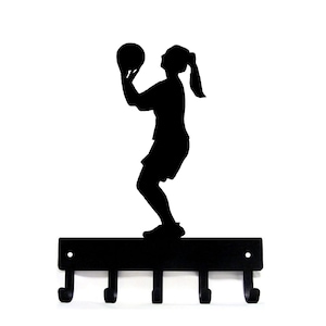 May include: Black metal wall hook featuring a silhouette of a basketball player in action. The player is poised to shoot the ball. Below the figure are four hooks for hanging items. The design is suitable for sports enthusiasts.