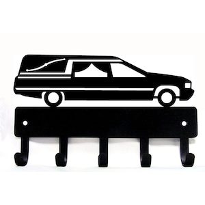 May include: Black metal key holder shaped like a hearse with five hooks. The hearse has a white interior and a white roof.