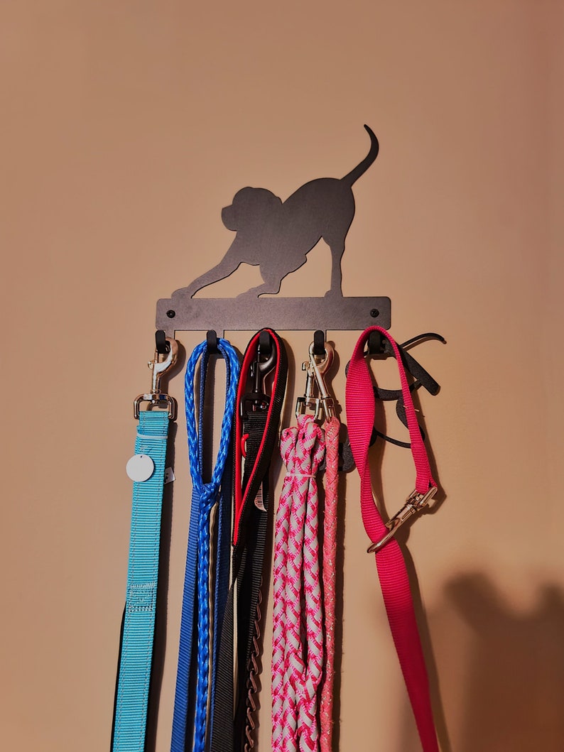 Black powder coated, five hook, key rack with the silhouette of a dog bowed in a playful stance with it's hind quarters and tail in the air. The rack is mounted on a tan wall and the hooks of the key rack hold multiple dog leashes.