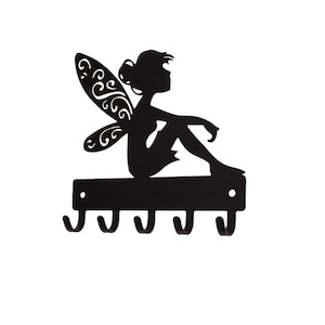 May include: Black metal key holder featuring a silhouette of a fairy with detailed wings, seated above four hooks. The fairy is in a relaxed pose, adding a whimsical touch to the functional design. Perfect for organizing keys or small items.