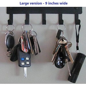 K&ouml;nnte beinhalten: Ein schwarzer Schl&uuml;sselhalter aus Metall mit f&uuml;nf Haken, an denen jeweils ein Schl&uuml;sselsatz und andere Gegenst&auml;nde h&auml;ngen. Der Halter ist 23 cm breit und an einer Wand montiert. Zu den Gegenst&auml;nden, die an den Haken h&auml;ngen, geh&ouml;ren ein Schl&uuml;sselanh&auml;nger, ein Autoschl&uuml;ssel, eine Taschenlampe und ein Schl&uuml;sselsatz mit verschiedenen Schl&uuml;sselanh&auml;ngern.