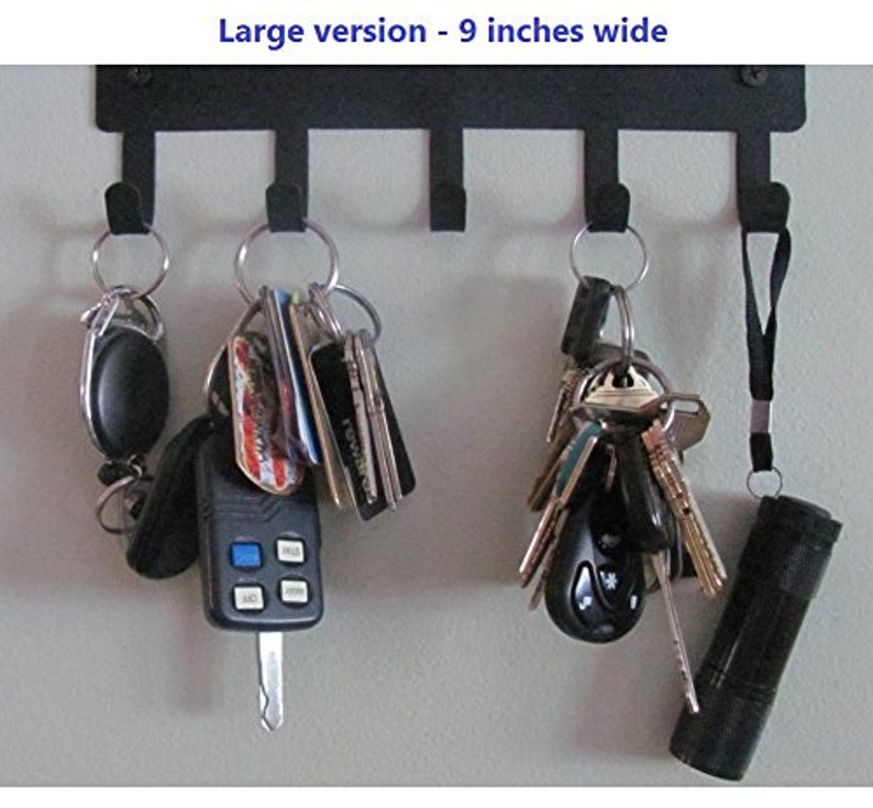 K&ouml;nnte beinhalten: Ein schwarzer Schl&uuml;sselhalter aus Metall mit f&uuml;nf Haken, an denen jeweils ein Schl&uuml;sselsatz und andere Gegenst&auml;nde h&auml;ngen. Der Halter ist 23 cm breit und an einer Wand montiert. Zu den Gegenst&auml;nden, die an den Haken h&auml;ngen, geh&ouml;ren ein Schl&uuml;sselanh&auml;nger, ein Autoschl&uuml;ssel, eine Taschenlampe und ein Schl&uuml;sselsatz mit verschiedenen Schl&uuml;sselanh&auml;ngern.
