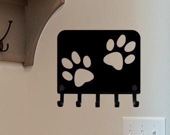 May include: Black metal key holder with five hooks and two white paw prints. The key holder is mounted on a wall.