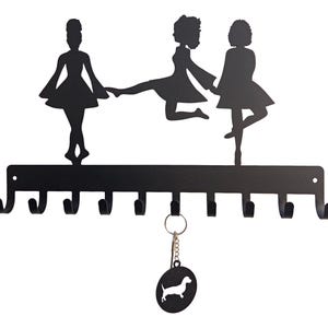 May include: Black metal wall-mounted key holder featuring silhouettes of three dancing figures above eight hooks. A dachshund keychain hangs below. The key holder is designed for organizing keys and adding a decorative touch to any space.
