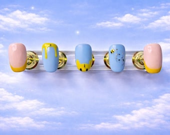 May include: Five artificial nails with a sky blue background. The nails are painted in various designs: pink with yellow tips, light blue with yellow drips, light blue with a yellow bee, and light blue with a bee design. The nails are mounted on a clear holder.