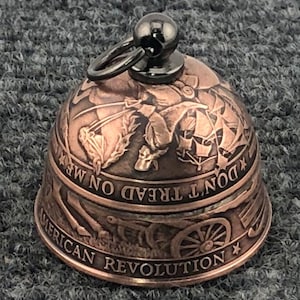 May include: A bronze-colored bell with a black loop and top. The bell features engraved imagery of the American Revolution, including soldiers, ships, and cannons. The words "DON'T TREAD ON ME" and "AMERICAN REVOLUTION" are also engraved.