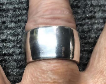 May include: A wide, polished silver ring. The ring is smooth and reflective, with a rounded band. The ring is shown on a finger, with the skin tone visible in the background. The ring appears to be made of metal.