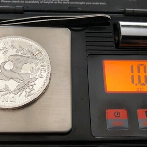 Puede incluir: Una moneda de plata con un dise&ntilde;o de &aacute;guila est&aacute; sobre una balanza digital, mostrando 28,58 gramos. La balanza negra tiene una pantalla naranja. Tambi&eacute;n se ven una peque&ntilde;a pesa de metal y unas pinzas.