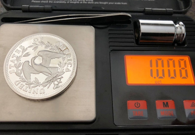 Puede incluir: Una moneda de plata con un dise&ntilde;o de &aacute;guila est&aacute; sobre una balanza digital, mostrando 28,58 gramos. La balanza negra tiene una pantalla naranja. Tambi&eacute;n se ven una peque&ntilde;a pesa de metal y unas pinzas.