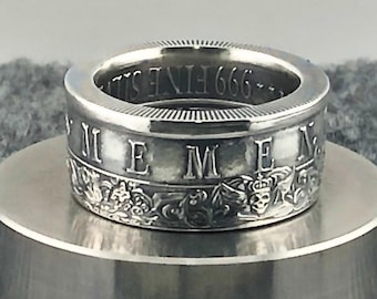 May include: A silver ring with the word "MEN" engraved on the side, along with decorative floral and skull motifs. The inner band of the ring is inscribed with "999 FINE SILVER". The ring is displayed on a silver surface.