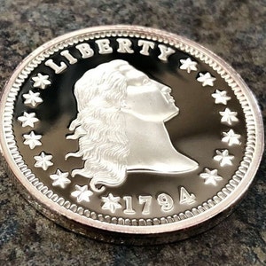 Puede incluir: Una moneda de plata con la palabra "LIBERTY" en la parte superior y el a&ntilde;o "1794" en la parte inferior. La moneda presenta un perfil de mujer con cabello suelto, rodeado de estrellas y un borde detallado.