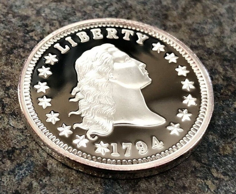 Puede incluir: Una moneda de plata con la palabra "LIBERTY" en la parte superior y el a&ntilde;o "1794" en la parte inferior. La moneda presenta un perfil de mujer con cabello suelto, rodeado de estrellas y un borde detallado.