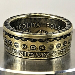 May include: A gold-toned metal ring crafted from a coin, featuring a serrated top edge and engraved text including "OSPOLITA POLSK", "KCYNORIK", and "ENIGMY 75". The band has a weathered, antique finish with dark recessed lettering and details.