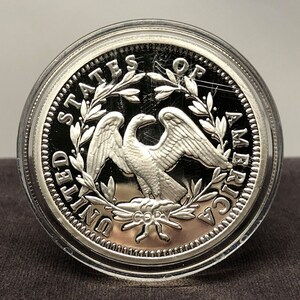 Puede incluir: Una moneda de plata con un &aacute;guila con las alas extendidas, rodeada por la inscripci&oacute;n "UNITED STATES OF AMERICA". La moneda est&aacute; dentro de una funda protectora transparente. La palabra "COPY" es visible.