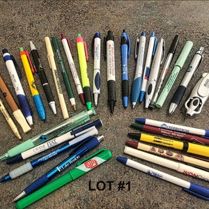 May include: A collection of 25 pens with various colors, logos, and designs. The pens are arranged in a semi-circle shape on a brown surface. The text "LOT #1" is visible in the center of the image.