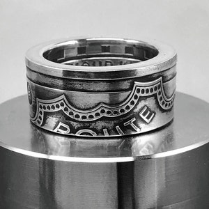 May include: A silver ring with the word "ROUTE" engraved on the side. The ring has a detailed, patterned design with a scalloped edge. The ring is displayed on a silver surface.
