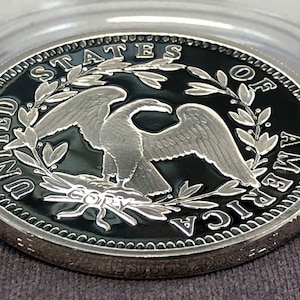 Puede incluir: Una moneda de plata con fondo negro, con un dise&ntilde;o de &aacute;guila y las palabras "UNITED STATES OF AMERICA". La moneda est&aacute; en una funda de pl&aacute;stico transparente. La palabra "COPY" es visible.