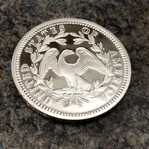 Puede incluir: Una moneda plateada con un dise&ntilde;o detallado de un &aacute;guila, rodeada por las palabras "UNITED STATES OF AMERICA". La moneda tiene un borde texturizado y una superficie brillante. La palabra "COPY" tambi&eacute;n es visible.