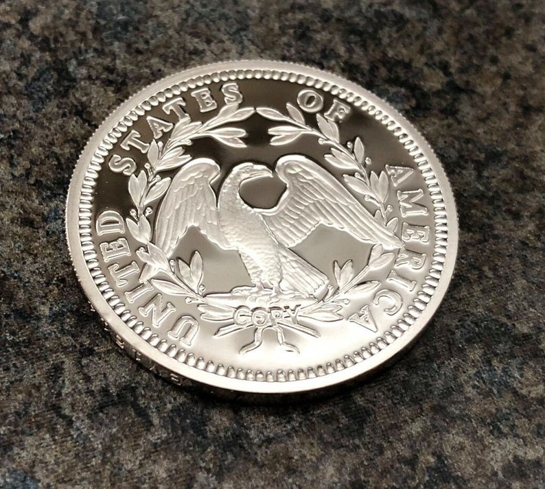 Puede incluir: Una moneda plateada con un dise&ntilde;o detallado de un &aacute;guila, rodeada por las palabras "UNITED STATES OF AMERICA". La moneda tiene un borde texturizado y una superficie brillante. La palabra "COPY" tambi&eacute;n es visible.