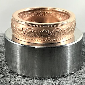 May include: A rose gold coin ring with intricate leaf and dot detailing. The ring features engraved text and a polished finish. The ring is displayed on a silver metal cylinder, showcasing the craftsmanship and design of the jewelry.
