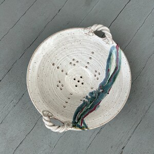 May include: White ceramic colander with two handles and a speckled glaze. The colander has a blue, green, and red glaze design on one side.