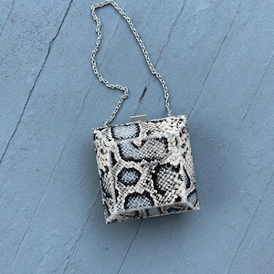 May include: A square-shaped handbag with a snakeskin pattern in black, white, and gray tones. The bag features a silver chain strap and a silver clasp closure. The bag is sitting on a blue wooden surface.