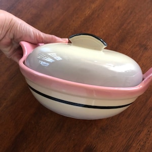 May include: A white and pink ceramic casserole dish with a black rim and a black handle on the lid. The dish has a black stripe around the rim.