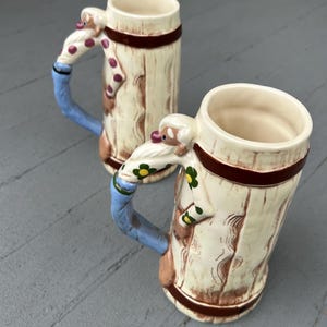 May include: Two ceramic beer steins with a whimsical design. The mugs have a light beige base with a wood grain texture, brown accents, and a red band. The handles are blue with a Santa Claus figure and a polka dot scarf.