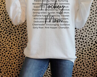 May include: White sweatshirt with black text that reads "Patient. Strong. Fierce. Nurturing. Warmhearted. Organised. Optimistic. Supportive. Hockey Mom. Passionate. Empowering. Protective. Strong-willed. Hustler. Mighty Mom. Caffeine Fueled. Game Day Traveller. Rink Enthusiast. Endless Constant. Dedicated Driver. Steady. Cheerleader. Encouraging. Guidance. Early Riser. Rink Hopper. Champion."