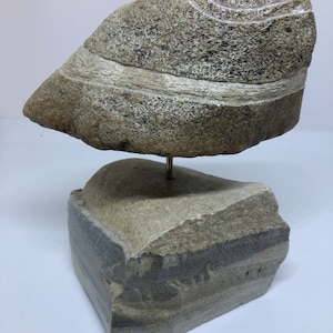 May include: A stone sculpture with a layered, striated design. The top piece is balanced on a smaller, rectangular base. Both pieces are a light brown color with hints of gray.