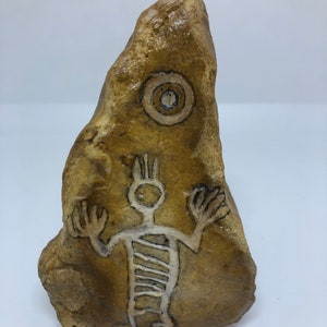 May include: A brown rock with a white drawing of a stick figure with raised arms and a spiral design on its torso. A white circle with a brown centre is drawn above the figure.