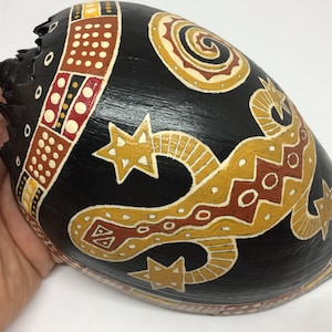 Könnte beinhalten: Eine schwarze Holz-Kalebasse mit einem bemalten Design eines Leguan, Sternen und geometrischen Mustern in Gelb, Rot und Weiß.