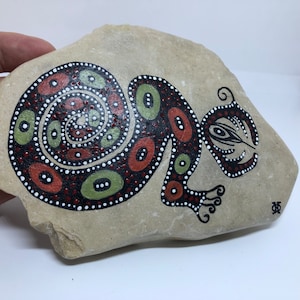 Peut inclure: Une pierre peinte à la main avec un motif de lézard stylisé en noir, vert, rouge et blanc. La queue du lézard est enroulée en spirale.