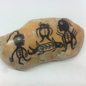 Könnte beinhalten: Ein handbemalter Stein mit schwarzer Tinte, der drei stilisierte Figuren und einen Fisch darstellt. Die Figuren haben große Augen und sind in einem einfachen, cartoonartigen Stil gezeichnet.