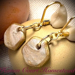 May include: Close-up of gold-colored leverback earrings with a geometric, stone-like pendant. The pendants have a gold-colored border and a light, pearlescent center. The earrings are displayed on a golden surface.