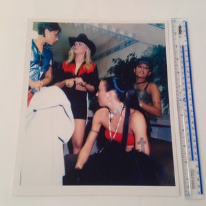 May include: Four women in a dimly lit room, one wearing a red top and a cowboy hat, another wearing a black top and a black hat, and two others wearing black tops. The woman in the red top is holding a microphone.