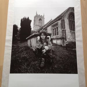 May include: A black and white photograph of a person sitting in front of a church. The person is wearing a jacket and has their arms crossed. The church is in the background and has a tall steeple.