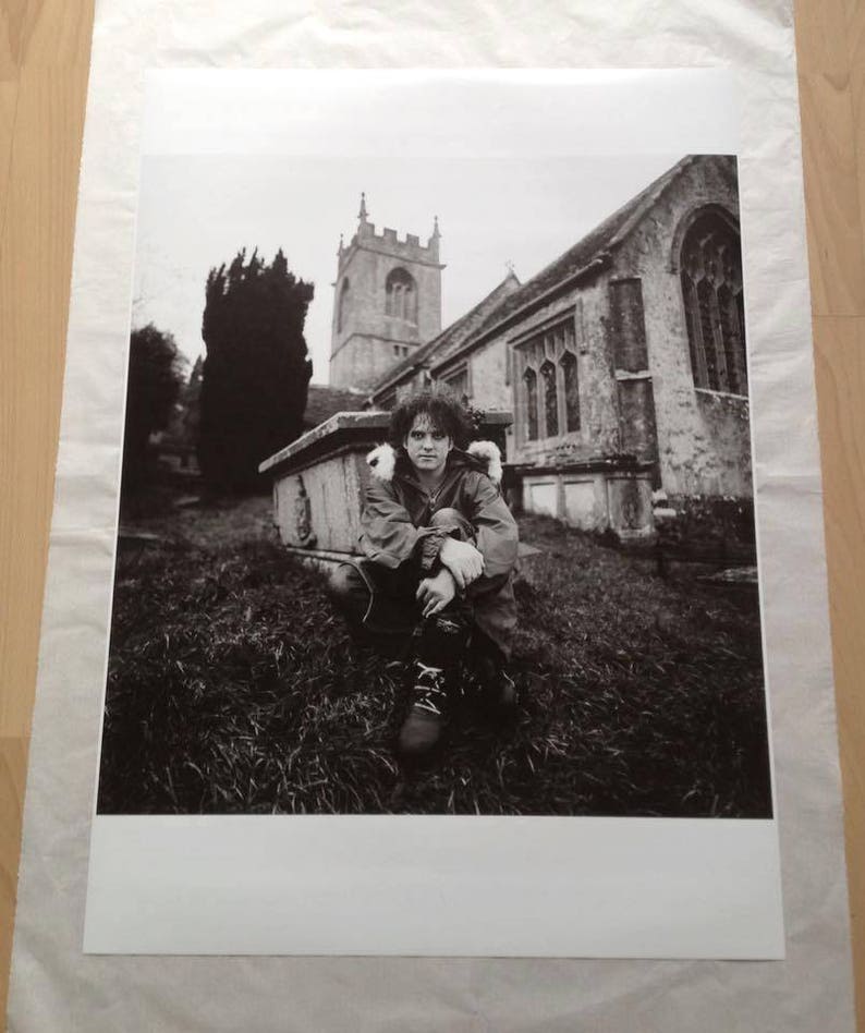 May include: A black and white photograph of a person sitting in front of a church. The person is wearing a jacket and has their arms crossed. The church is in the background and has a tall steeple.
