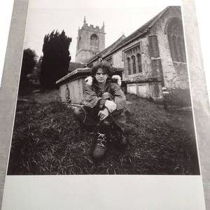 May include: A black and white photograph of a person sitting in front of a church. The person is wearing a jacket and has their arms crossed. The church is in the background and has a tall steeple.