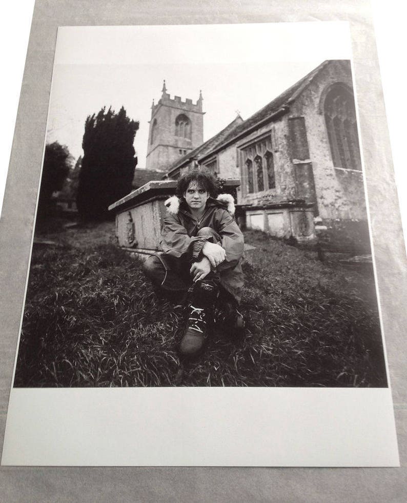 May include: A black and white photograph of a person sitting in front of a church. The person is wearing a jacket and has their arms crossed. The church is in the background and has a tall steeple.